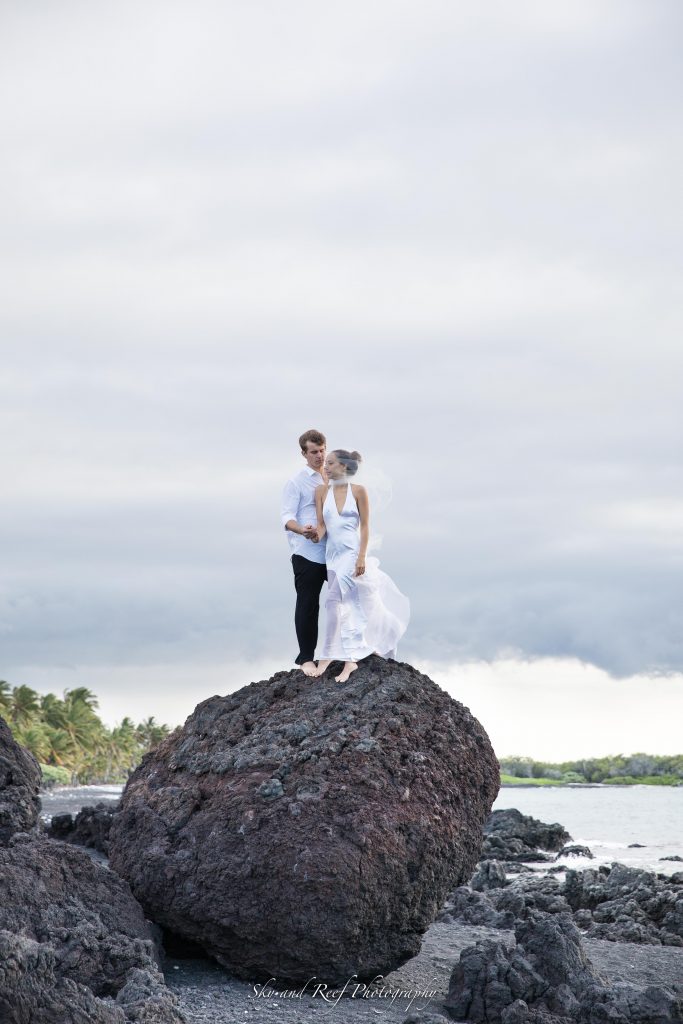 bride and groom stand atop a large rock on beach elopement